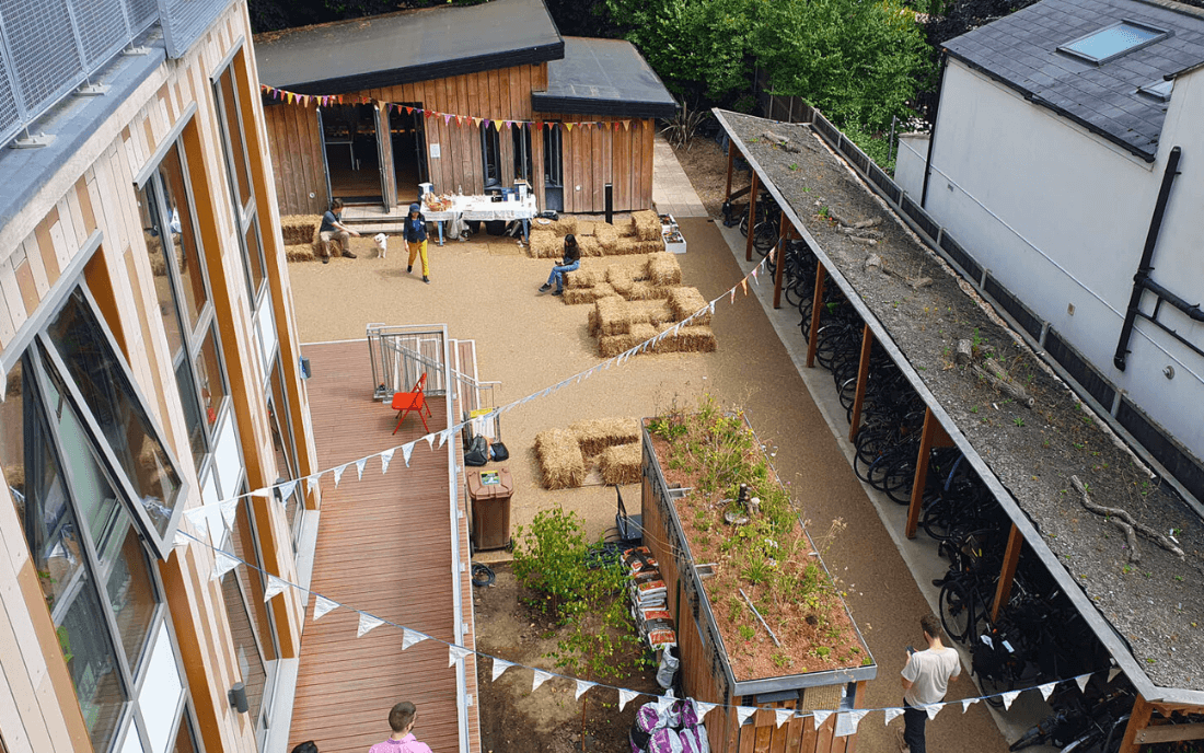 Church Grove A courtyard view of Church Grove housing development, showing wooden-clad buildings, communal seating with hay bales, bunting decorations, and residents gathered in a welcoming shared space.