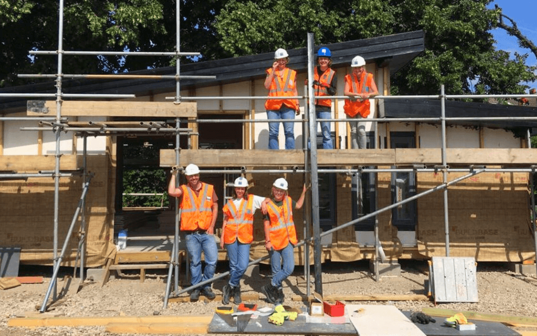 high-vis-building Construction workers wearing high-visibility vests and hard hats standing on scaffolding and in front of a timber-frame building under construction, surrounded by tools and safety equipment on a sunny day.